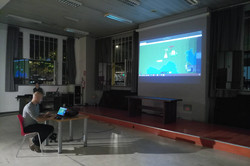 Ein Mensch sitzt allein an einem Tisch mit Laptop in einem Raum. Auf einer großen Leinwand wird ein buntes Bild projiziert. Der Raum ist schwach beleuchtet.