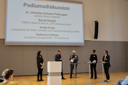 Foto: Mehrere Personen stehen auf einer Bühne während einer Podiumsdiskussion. Auf der Leinwand sind die Namen und Funktionen der Teilnehmenden aufgeführt, darunter das Landesamt für Natur, Umwelt und Verbraucherschutz NRW sowie die Sozialforschungsstelle der TU Dortmund.