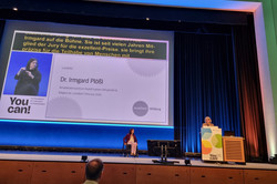 Foto: Eine Person steht auf einer Bühne hinter einem Rednerpult. Hinter ihr wird auf einer großen Leinwand eine Präsentation gezeigt. Vor der Bühne sitzen mehrere Personen.