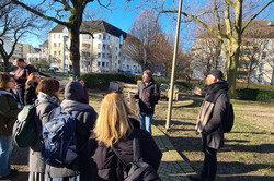 Eine Gruppe von Personen steht im Freien in einem städtischen Bereich mit Bäumen und Gebäuden. Eine Person spricht zu der Gruppe, während die anderen zuhören.