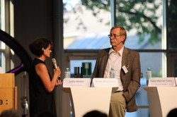 Eine Person spricht mit einem Mikrofon an einem Stehtisch zu einer weiteren Person. Auf den Tischen stehen Namensschilder, Wasserflaschen und Mikrofone.