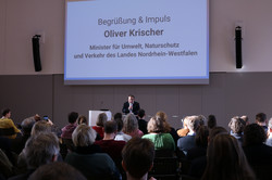 Oliver Krischer steht auf einer Bühne. Vor ihm sitzen viele Menschen und schauen ihn an. Auf einer Leinwand wird eine Präsentation angezeigt.