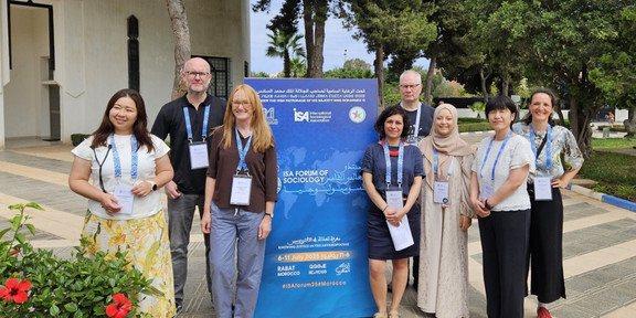 The eight participants in the panel discussion stand next to a display stand for the ISA conference.
