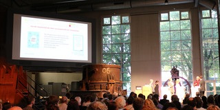 Foto: Publikum verfolgt eine Präsentation auf einer Bühne in einem Veranstaltungsraum mit großer Projektionsfläche.