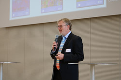 Eine Person spricht in ein Mikrofon, das sie in der Hand hält. Sie steht zwischen zwei Stehtischen. Hinter ihr wird ein Bild an eine Leinwand projiziert.