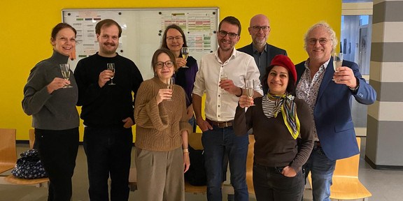 Eine Gruppe von Menschen steht in einem Raum mit gelber Wand und stößt mit Gläsern an.