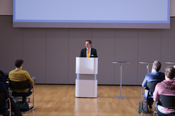 Eine Person steht an einem Rednerpult und spricht vor mehreren sitzenden Zuhörenden in einem Konferenzraum.