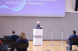Jürgen Schultze steht vor einem Rednerpult und hält ein Mikrofon in der Hand. Vor ihm sitzen vereinzelt Personen und schauen ihn und/oder eine Präsentation im Hintergrund an.