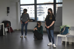 3 Personen sind in einem Raum. Zwei Personen stehen, eine hockt sich hin. Sie tragen Corona-Schutzmasken und halten Abstand zueinander.