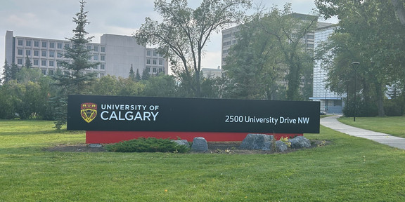 Photo: University of Calgary campus. Sign with the words “University of Calgary” and the address “2500 University Drive NW.” Several buildings and trees are visible in the background.
