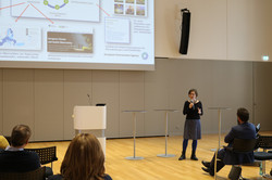 Eine Person steht auf einer Bühne in einem Saal und spricht vor Publikum. Im Hintergrund ist eine große Leinwand mit Präsentationsfolien.