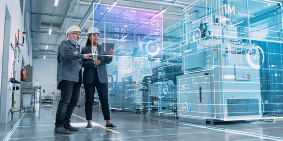 AdobeStock_555709923 Zwei Personen sehen sich in einer Fabrikhalle projizierte Leistungsdaten an.