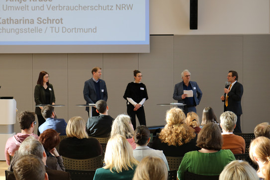 Podiumsdiskussion Foto: Podiumsdiskussion mit fünf Personen an Stehtischen und einem Moderator mit Mikrofon; Publikum sitzt in Reihen und hört zu.