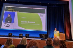 Foto: Eine Person steht auf einer Bühne hinter einem Rednerpult. Hinter ihr wird eine Präsentation auf einer großen Leinwand angezeigt. Vor der Bühne sitzen mehrere Personen.