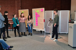 Foto: Kleine Gruppe in einer Workshop-Situation; mehrere Teilnehmende stehen an den Pinnwänden, ein Präsentierender mit Laptop spricht, weitere Teilnehmende sitzen an der Seite.