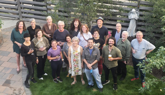 Photo: A group of 18 people are standing in a garden area. Some are holding drinks in their hands. Plants and a statue are visible in the background.
