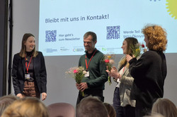 Foto: Vier Personen stehen vor einem Publikum, drei von ihnen halten einen Blumenstrauß in der Hand.