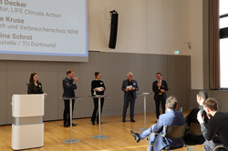 5 Personen stehen vor einer Zuhörerschaft. Vor ihnen sind ein Rednerpult und drei Stehtische aufgebaut. Hinter ihnen wird eine Präsentation auf eine Leinwand projiziert.