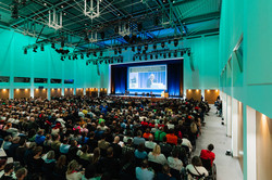 Foto: Viele Personen sitzen in mehreren Stuhlreihen in einem Saal. Auf einer Bühne wird eine Präsentation angezeigt.