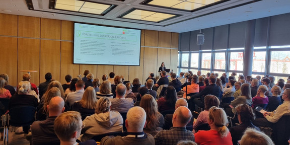 Lisa Preissner steht in einem Saal, in dem viele Personen in mehreren Reihen auf Stühlen sitzen. Sie steht an einem Rednerpult und hat einen aufgeklappten Laptop vor sich. Auf einer Leinwand wird eine Präsentation gezeigt.