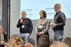 Foto: Vier Personen vor einem Publikum, drei stehen, eine Person sitzt. Sie halten Mikrofone in der Hand und tragen ID-Karten um ihren Hals.