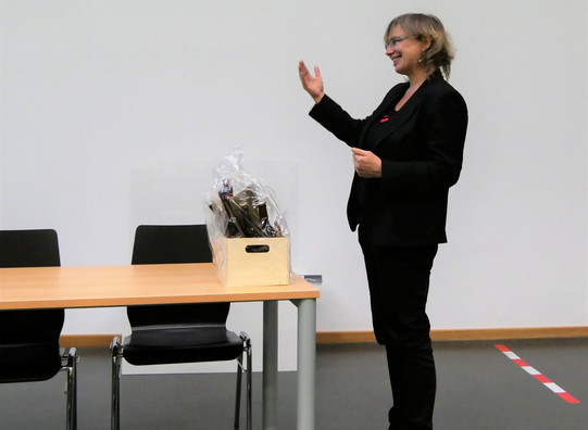  The photo shows Prof. Nicole Burzan, Dean of the Department of Social Sciences. She is standing at a table in the front of the lecture hall during Prof. Mona Motakef's inaugural lecture. In front of her is a small gift basket.