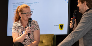 Prof. Dr. Martina Brandt im Gespräch mit Prof. Dr. Michael Steinbrecher Prof. Dr. Martina Brandt sitzt auf der linken Seite. Sie unterhält sich Prof. Dr. Michael Steinbrecher, der rechts sitzt. Beide halten ein Mirkofon in der Hand.