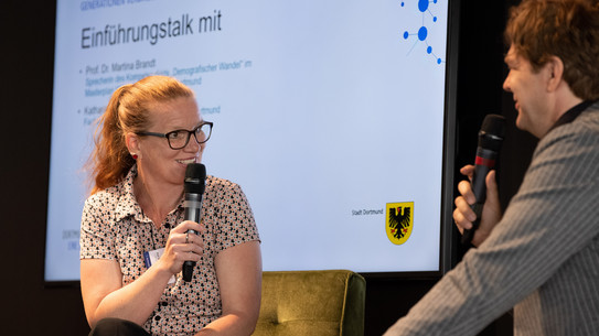 Prof. Dr. Martina Brandt sitzt auf der linken Seite. Sie unterhält sich Prof. Dr. Michael Steinbrecher, der rechts sitzt. Beide halten ein Mirkofon in der Hand.