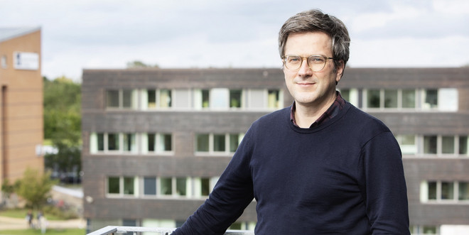 Bei dem Foto handelt es sich um eine Porträt von Prof. Dr. Bernd Sommer. Er steht auf einem Balkon und hält sich mit der rachten Hanf am Geländer fest. Im Hintergrund ist ein Gebäude zu sehen.