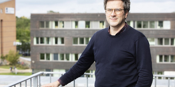 The photo is a portrait of Prof. Bernd Sommer. He is standing on a balcony and holding onto the railing with his right hand. In the background you can see a building.