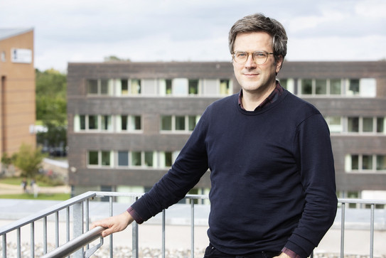 Bei dem Foto handelt es sich um eine Porträt von Prof. Dr. Bernd Sommer. Er steht auf einem Balkon und hält sich mit der rachten Hanf am Geländer fest. Im Hintergrund ist ein Gebäude zu sehen.