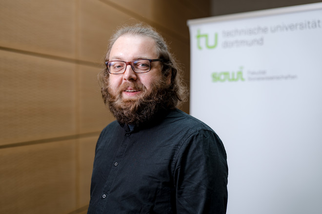  The photo shows a portrait of Dr. Paul Eisewicht. Behind him is a roll-up displaying the logos of the Technical University of Dortmund and the Department of Social Sciences.