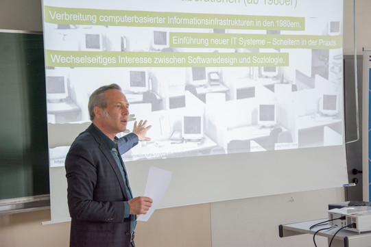 Auf dem Foto steht links Prof. Dr. Cornelius Schubert, der einen Vortrag hält. Im Hintergrund ist eine Leinwand mit einer Präsentation zu sehen, auf die Prof. Dr. Schubert zeigt.