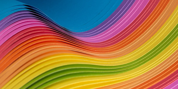 The abstract image consists of thin fabrics layered on top of each other. They form a wave with a colorful gradient of blue at the upper left and lower right edge over pink, red, orange, yellow and green in the center of the picture.