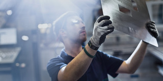 A worker wears gloves and lifts a large metal sheet with rectangular openings cut out against the light in a factory hall.