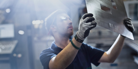 A worker wears gloves and lifts a large metal sheet with rectangular openings cut out against the light in a factory hall.
