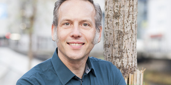Das Foto zeigt ein Protrait von Prof Dr. Cornelius Schubert, der draußen vor einem Baum am Straßenrand steht.
