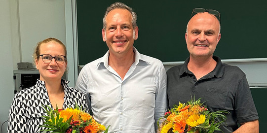 Auf dem Foto ist das neu gewählte Dekanat der Fakultät Sozialwissenschaften zu sehen. Links steht die wiedergewählte Prodekanin Forschung, Prof. Dr. Martina Brandt, mit einem Blumenstrauß in der Hand. In der Mitte steht der neugewählte Dekan, Prof. Dr. Cornelius Schubert. Rechts neben ihm steht der wiedergewählte Studiendekan, Dr. Arne Niederbacher, der ebenfalls einen Blumenstrauß in der Hand hält.