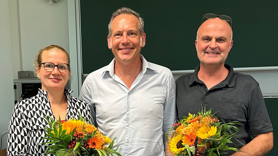 The photo shows the newly elected Dean's Office of the Department of Social Sciences. On the left is the re-elected Vice Dean of Research, Prof. Martina Brandt, with a bouquet of flowers in her hand. In the middle is the newly elected Dean, Prof. Cornelius Schubert. To his right is the re-elected Dean of Studies, Dr. Arne Niederbacher, who is also holding a bouquet of flowers.