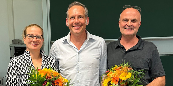 The photo shows the newly elected Dean's Office of the Department of Social Sciences. On the left is the re-elected Vice Dean of Research, Prof. Martina Brandt, with a bouquet of flowers in her hand. In the middle is the newly elected Dean, Prof. Cornelius Schubert. To his right is the re-elected Dean of Studies, Dr. Arne Niederbacher, who is also holding a bouquet of flowers.