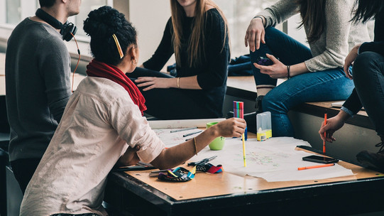 In the center of the picture is a table on which many papers are spread out. A group of students sits around the table and discusses.