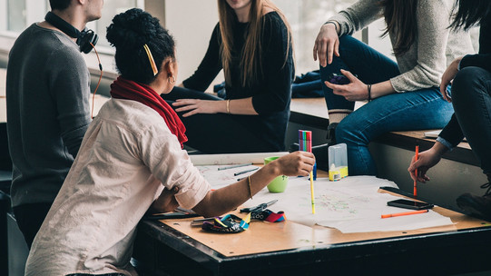Im Zentrum des Bildes ist ein Tisch auf dem viele Papiere ausgebreitet sind. Eine Gruppe studierenden sitzt um den Tisch herum und diskutiert.