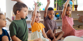 Mehrere Kinder sitzen in einem Klassen- oder Gruppenraum und heben die Hand, teilweise mit zwei ausgestreckten Fingern zum Abstimmen.
