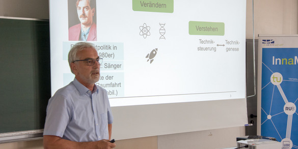 On the left side of the picture is Prof. Weyer, who is giving a lecture. In the background, a presentation can be seen on a screen.