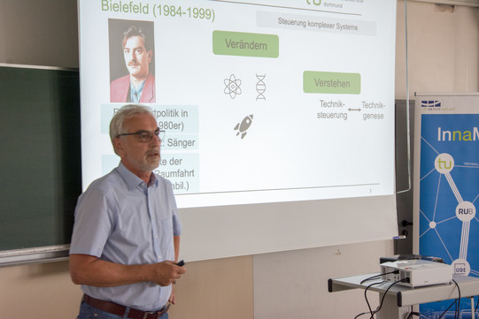 Auf der linken Seite des Bildes steht Prof. Dr. Weyer, der einen Vortrag hält. Im Intergrund ist auf einer Leinwand eine Präsentation zu sehen.