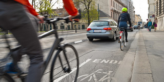 Abgebildet ist eine von zahlreichen Gebäuden umgebene Verkehrsstraße, in der Radfahrer im Mittelpunkt stehen. Ein Radfahrer im Vordergrund ist verschwommen dargestellt und fährt schnell, während eine zweite Person auf einem Fahrrad auf einer markierten Spur hinter einem silbernen Auto fährt.