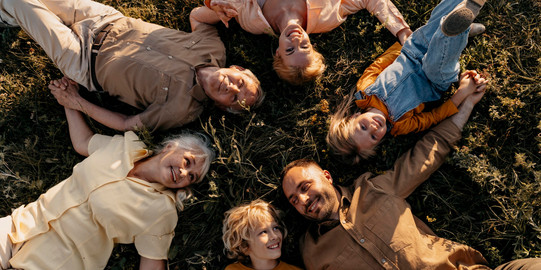 Eine Menschengruppe verschiedenen Alters liegt im Kreis lachend im Gras.