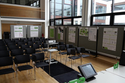 Fakultätsforschungstag 2024_Raumansicht Auf dem Foto sind ist der menschenleere Raum des Fakultätsforschungstages vor Beginn der Veranstaltung zu sehen. In der Mitte sind Stuhlreihen aufgebaut. Außen an den Wänden sind Stellwände mit den Postern positioniert.