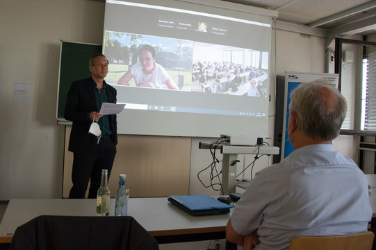 Auf dem Foto steht links Prof. Dr. Cornelius Schubert. Rechts vorne sitzt Prof. Dr. Johannes Weyer mit dem Rücken zur Kamera. Im Hintergrund ist eine Leinwand zu sehen. Rechts auf der Leinwand ist Frau Prof. Dr. Cornelia Kropp zu sehen, die digital zugeschaltet ist. Auf der rechten Seite der Leinwand, sieht man die Kameraaufnahme des Auditoriums.