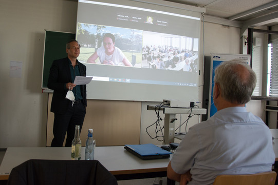 On the left in the photo is Prof. Cornelius Schubert. Prof. Johannes Weyer is sitting in front on the right with his back to the camera. A screen can be seen in the background. On the right side of the screen, you can see Prof. Cornelia Kropp, who is digitally connected. On the right side of the screen, you can see the camera shot of the auditorium.